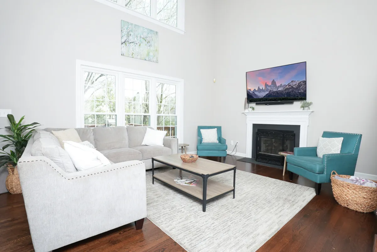 The hero image features a spacious and stylish living room with a plush gray sectional sofa, vibrant teal accent chairs, a sleek modern coffee table, and a cozy fireplace topped with a large TV. Sunlight pours in through expansive windows, creating an inviting atmosphere ideal for relaxing, entertaining, or family movie nights.