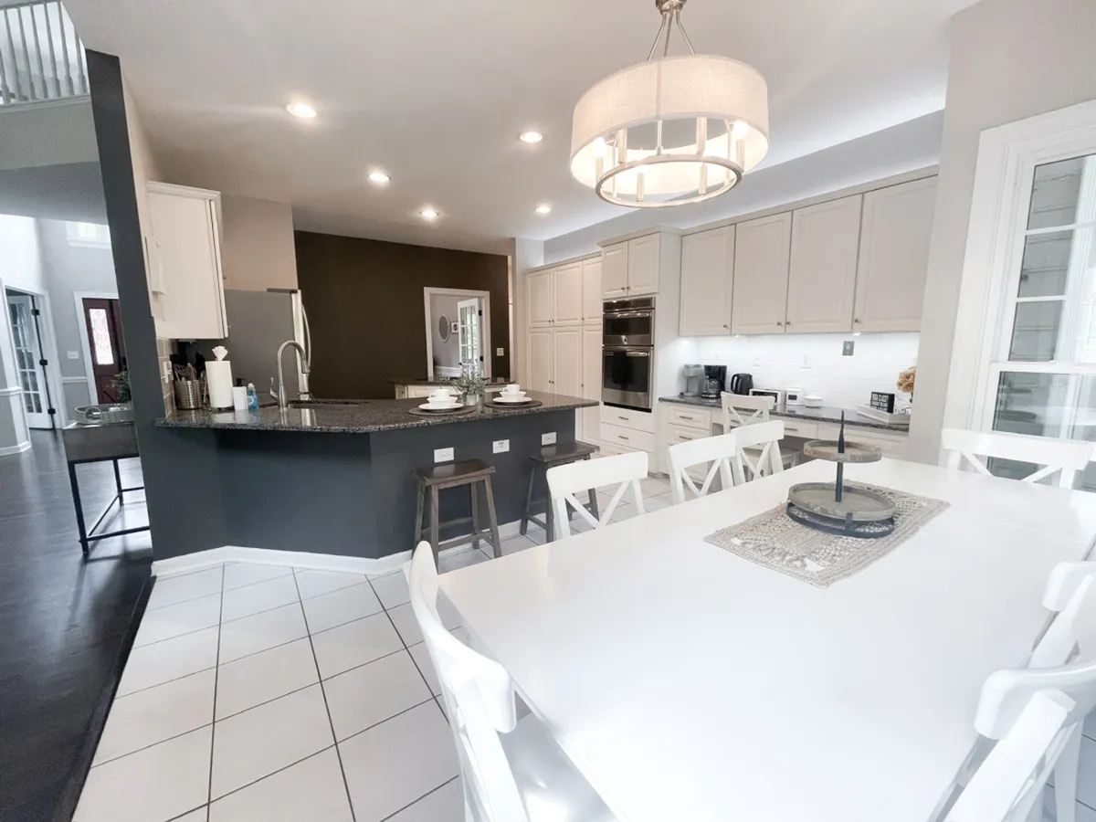 A modern interior view featuring a large dining table, a central island, an elegant chandelier, and visible kitchen appliances. The space is clearly designed for both cooking and communal dining, evoking a sense of celebration and togetherness.