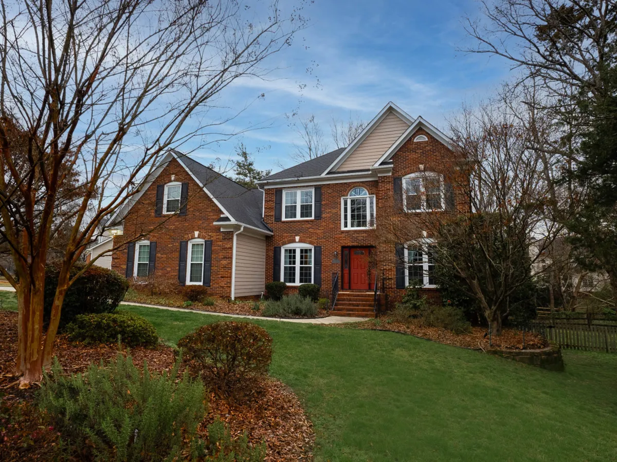 A welcoming brick home with a striking red front door, manicured lawn, and mature trees, providing an inviting entrance and plenty of room for outdoor play or relaxation.