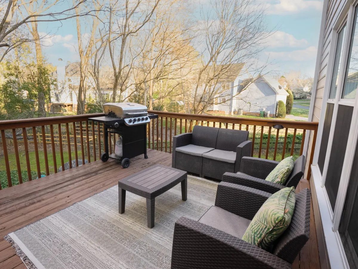A spacious back deck is furnished with a BBQ grill, outdoor seating, and a decorative rug. The deck is surrounded by lush woods, creating a private and relaxing atmosphere ideal for al fresco dining, grilling, and socializing at any time of day, with views that invite connection to nature.