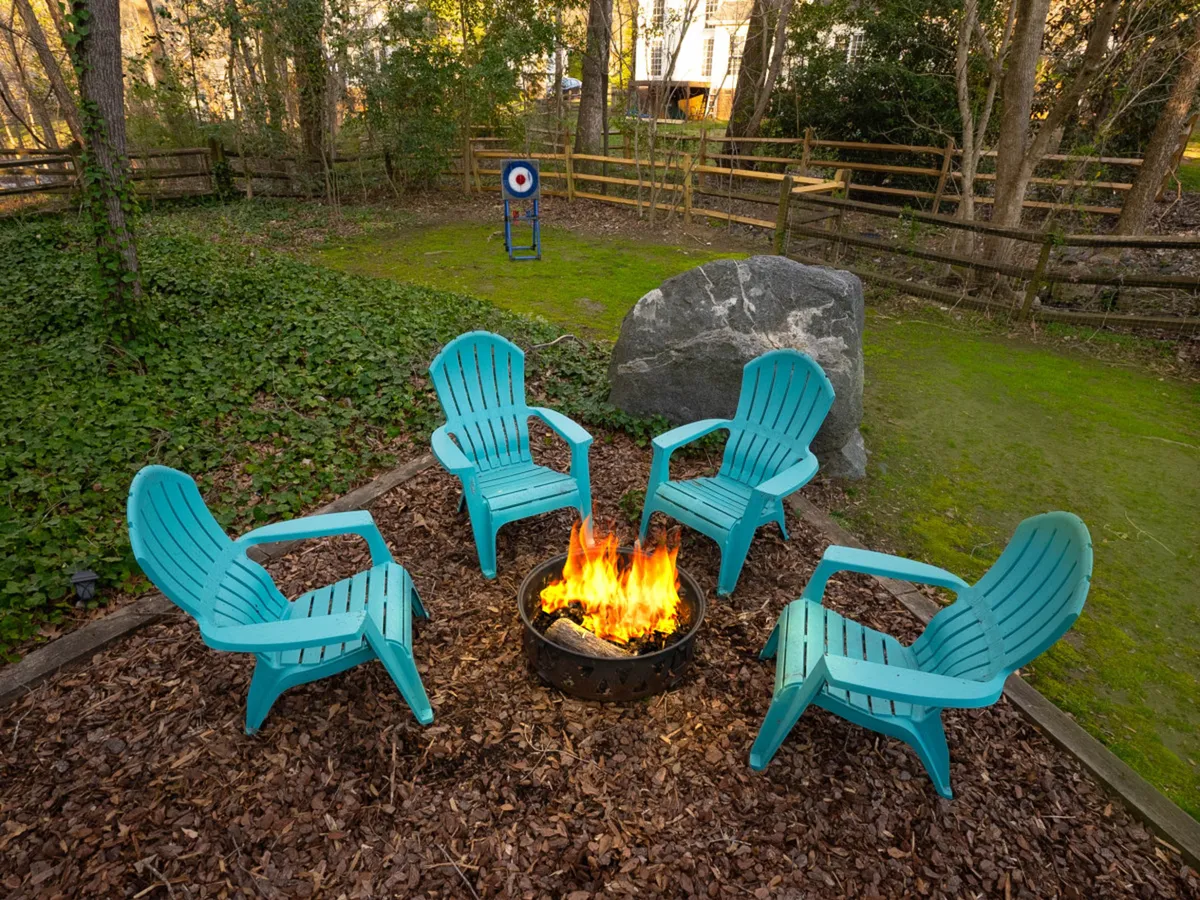 A cozy fire pit scene features turquoise chairs arranged in a circle, a large rock, and hints of playful activities like a target in the background. The space is clearly designed for gathering, storytelling, and roasting marshmallows, with woodland surroundings enhancing the sense of retreat and community.