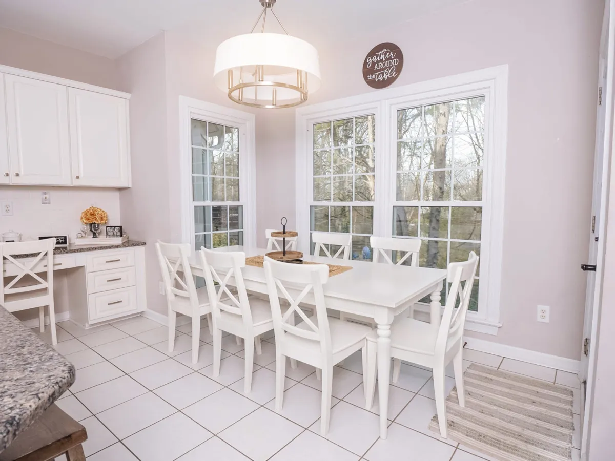 A cozy dining area featuring a large wooden table, elegant chandelier, and expansive windows that bathe the space in natural light. The arrangement suggests a welcoming setting for family meals, celebrations, and casual gatherings, set within sight of the kitchen.