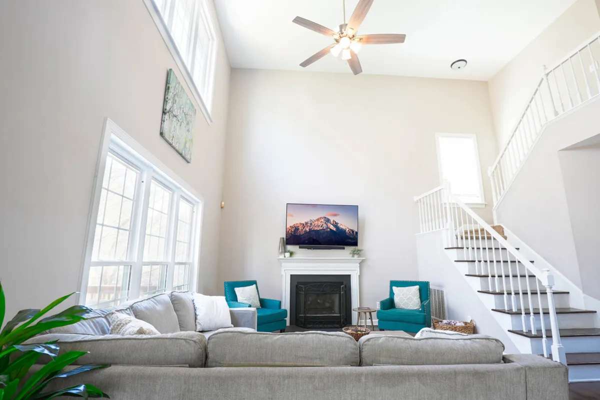 A spacious living room featuring a large plush sectional sofa arranged around a mounted television above a modern fireplace, with green accent chairs and a sweeping staircase leading to upper levels. Floor-to-ceiling windows allow natural light to flood the space, creating an inviting atmosphere for family gatherings or movie nights.