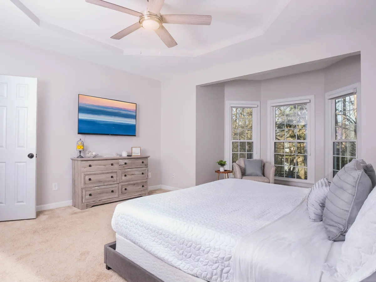 A serene bedroom with a comfortable bed, dresser, windows letting in natural light, a chair for reading, and artwork on the walls. The atmosphere is peaceful and inviting, perfect for relaxation and restful nights.