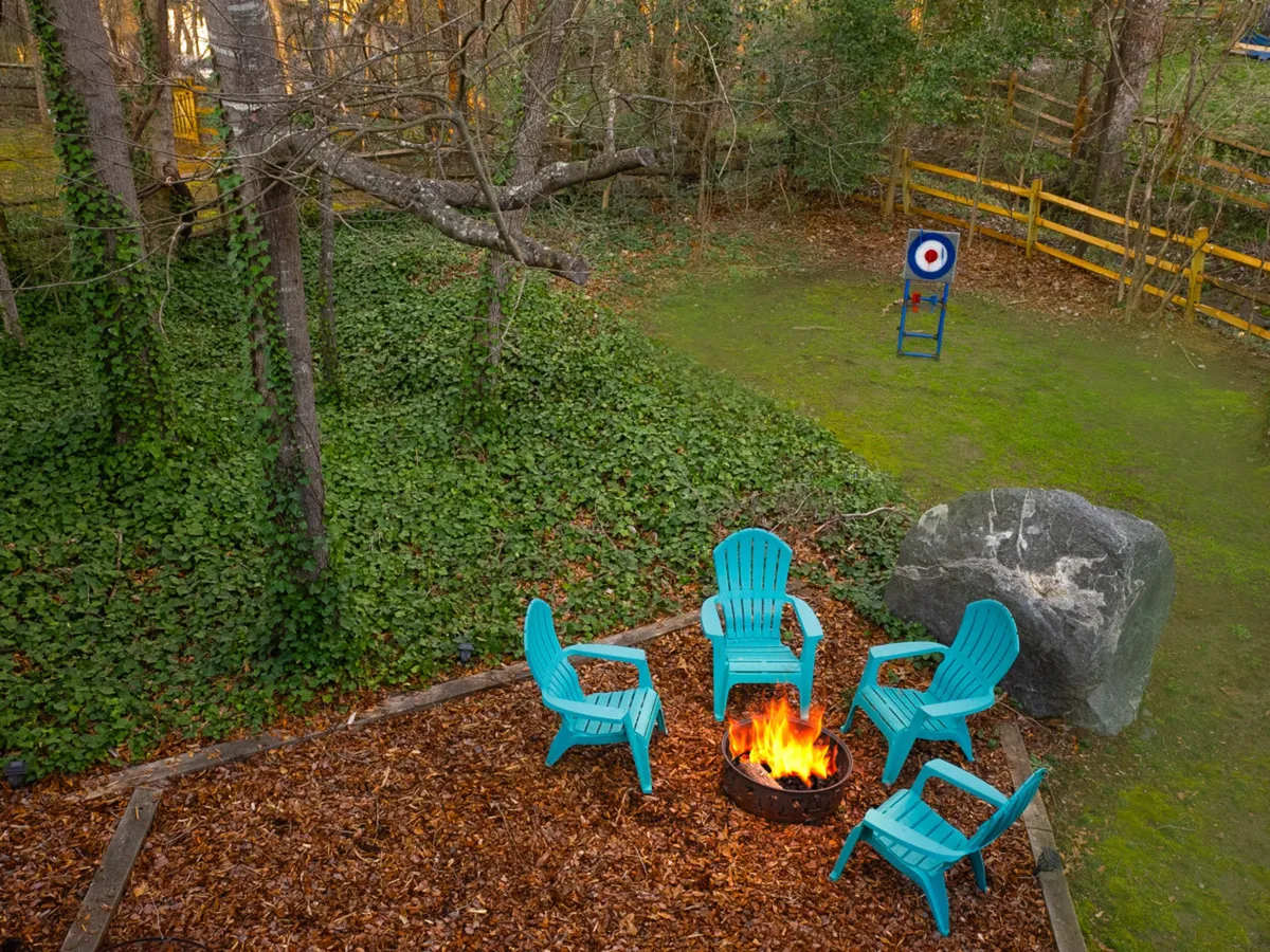 A cozy backyard scene with a fire pit surrounded by turquoise chairs, a large rock, and archery target, all set within lush greenery. The setup invites evening gatherings and outdoor adventures like roasting marshmallows or light games.