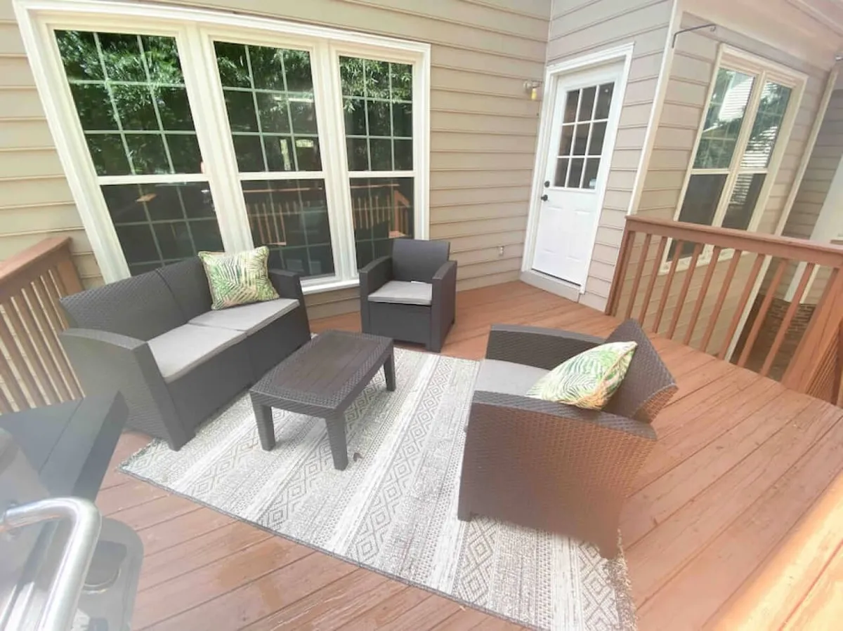 This exterior photo features a cozy wicker furniture set arranged on a deck, adorned with decorative cushions and a rug. Large windows connect the outdoor seating to the home’s interior, while the surrounding greenery hints at a wooded, private atmosphere. The scene is ideal for sipping coffee in the morning or enjoying intimate conversations under the stars.